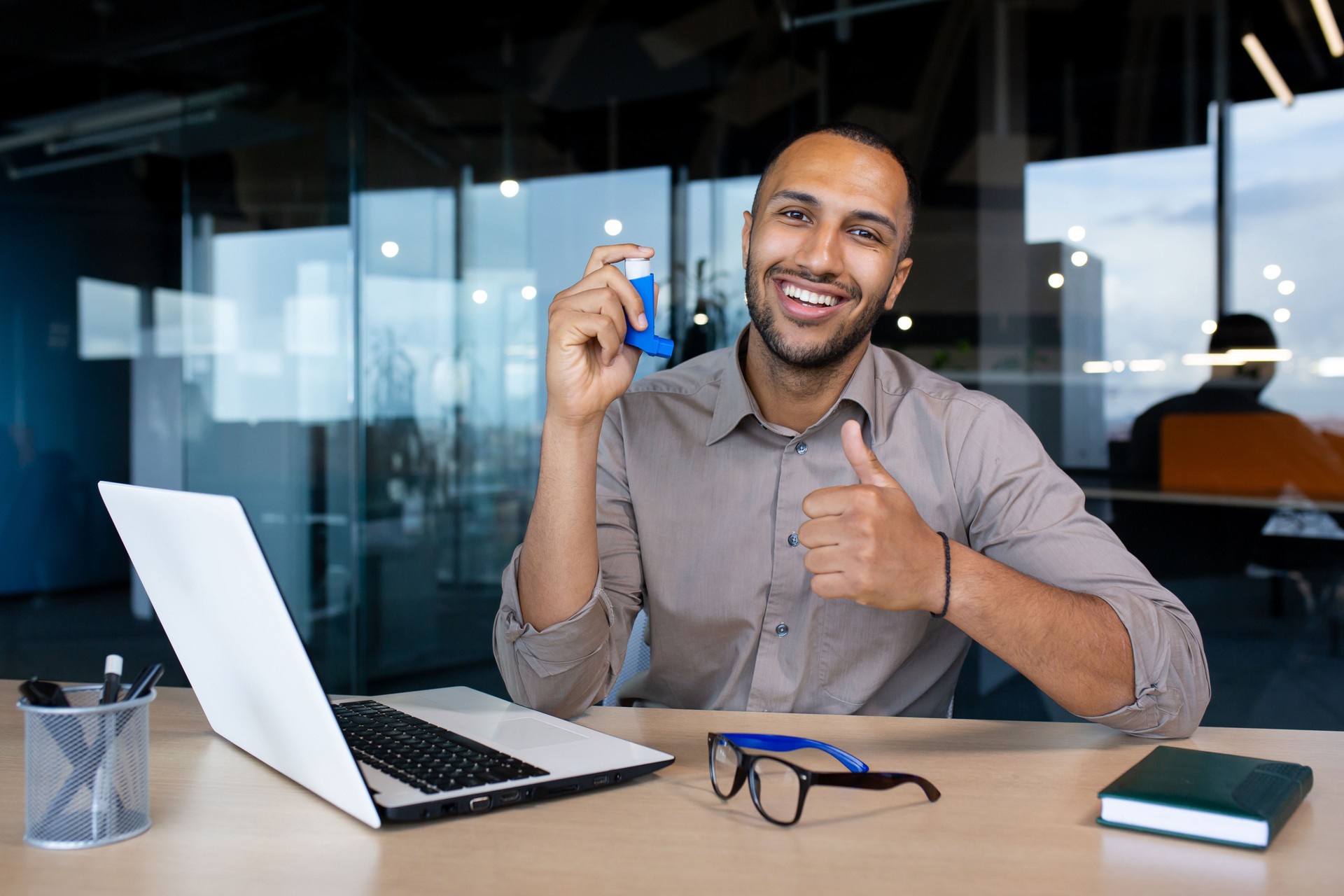 African american guy sitting in office with asthma inhaler in hand, showing thumbs up like, smiling and looking at camera